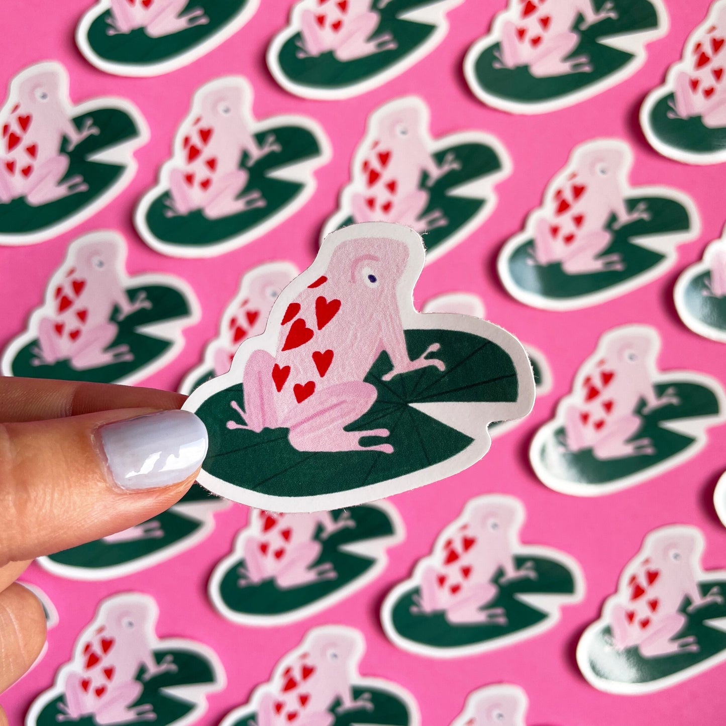 small vinyl sticker with a pink frog with hearts on it sitting on a dark green lily pad 