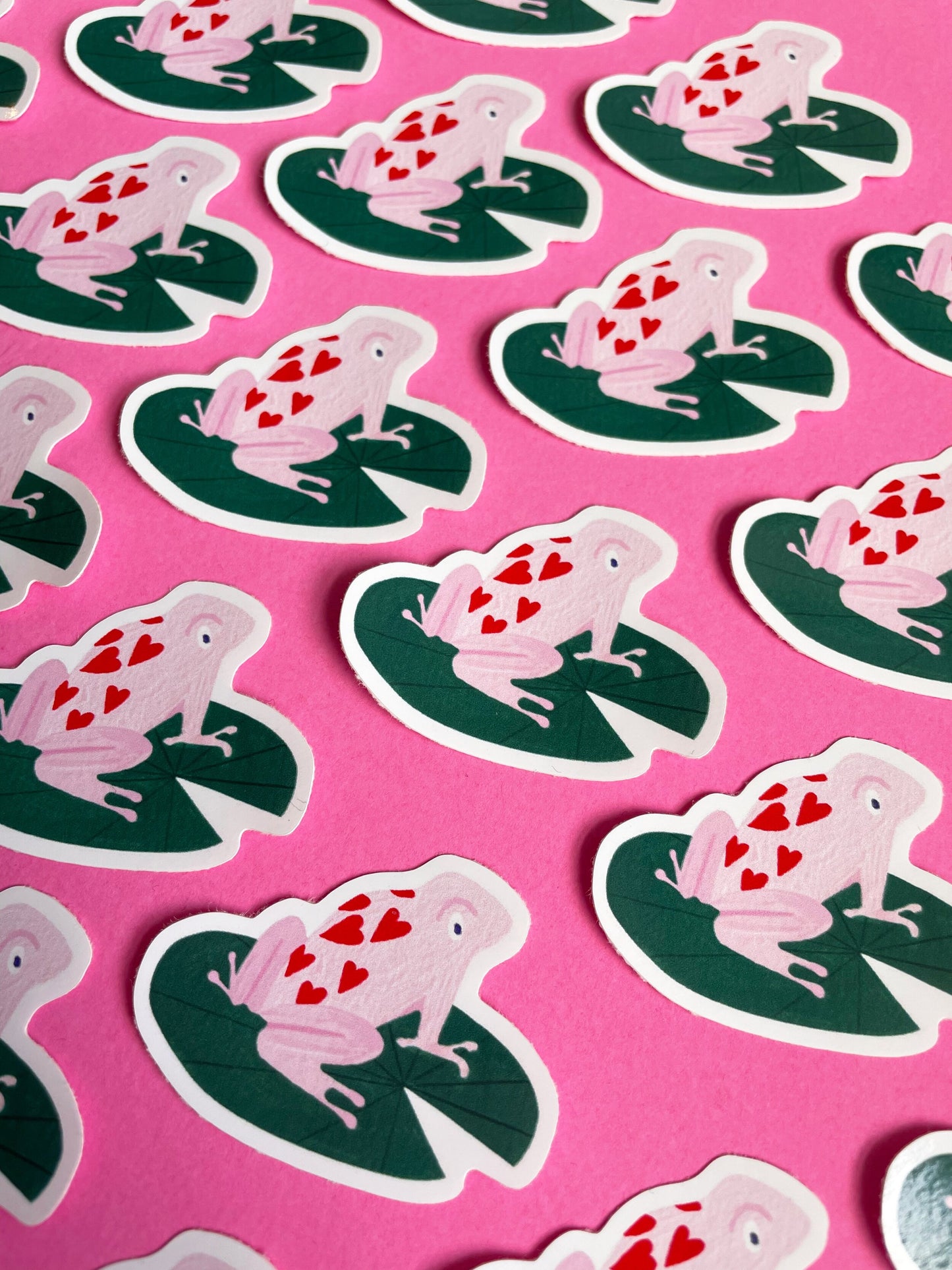 small vinyl sticker with a pink frog with hearts on it sitting on a dark green lily pad