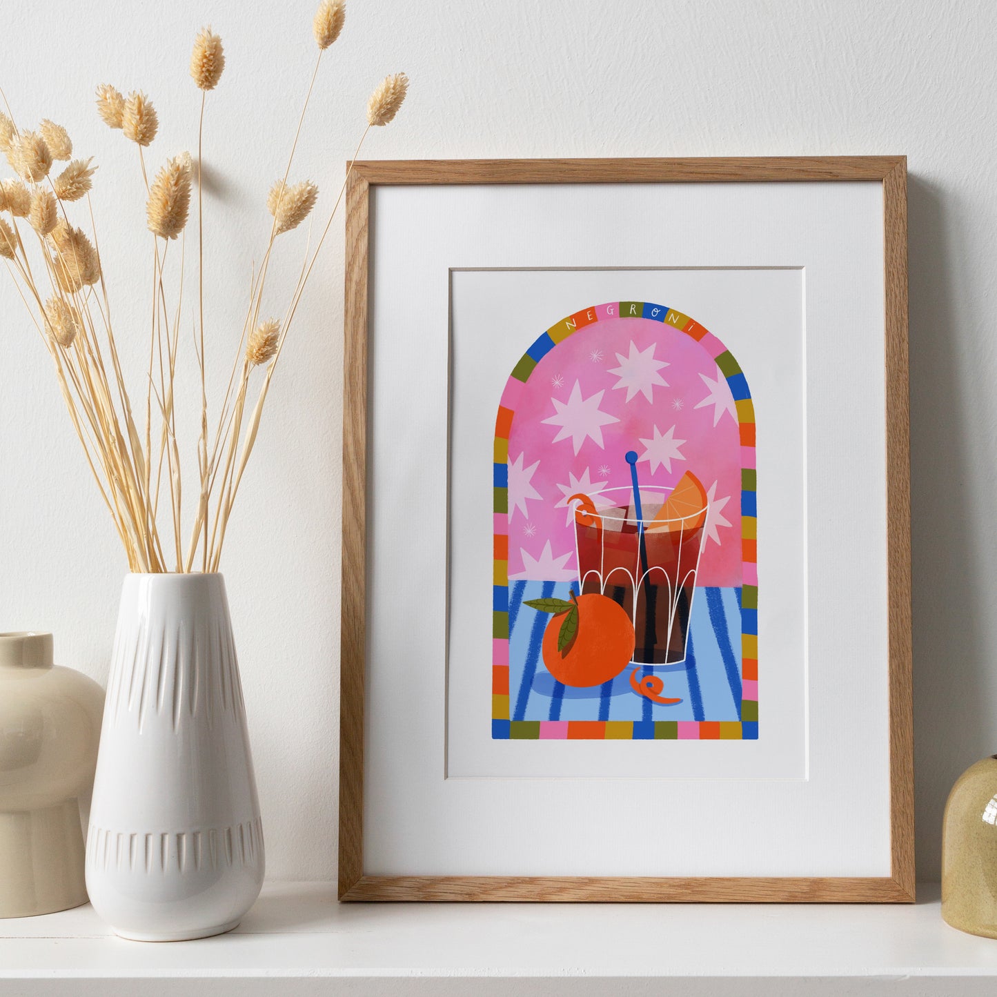 fun colourful art print of a negroni cocktail with an orange in the foreground and orange peel on the blue stripy table. The background is pink with stars and a rainbow arched border reads negroni at the top
