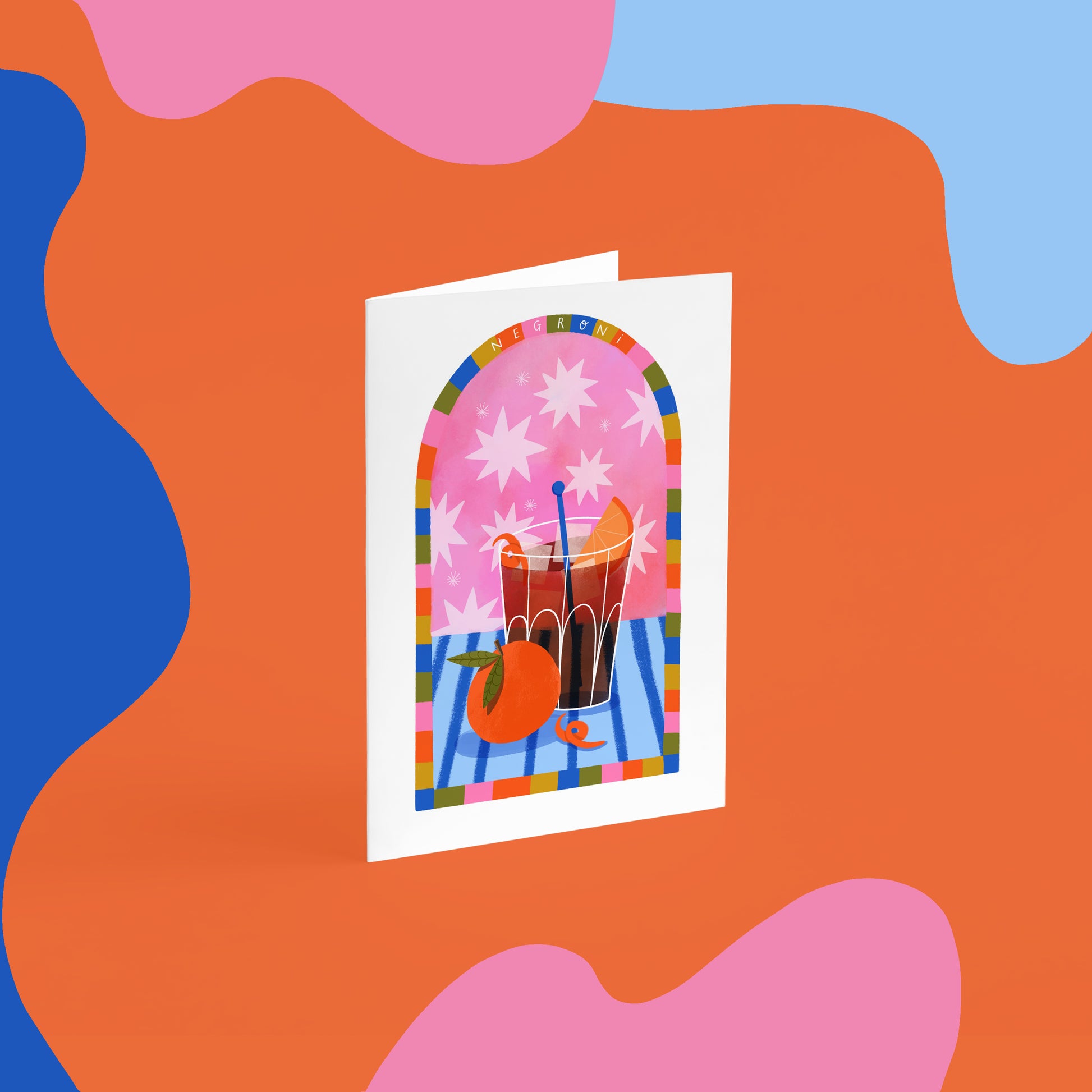 a6 greetings card. fun colourful art print of a negroni cocktail with an orange in the foreground and orange peel on the blue stripy table. The background is pink with stars and a rainbow arched border reads negroni at the top