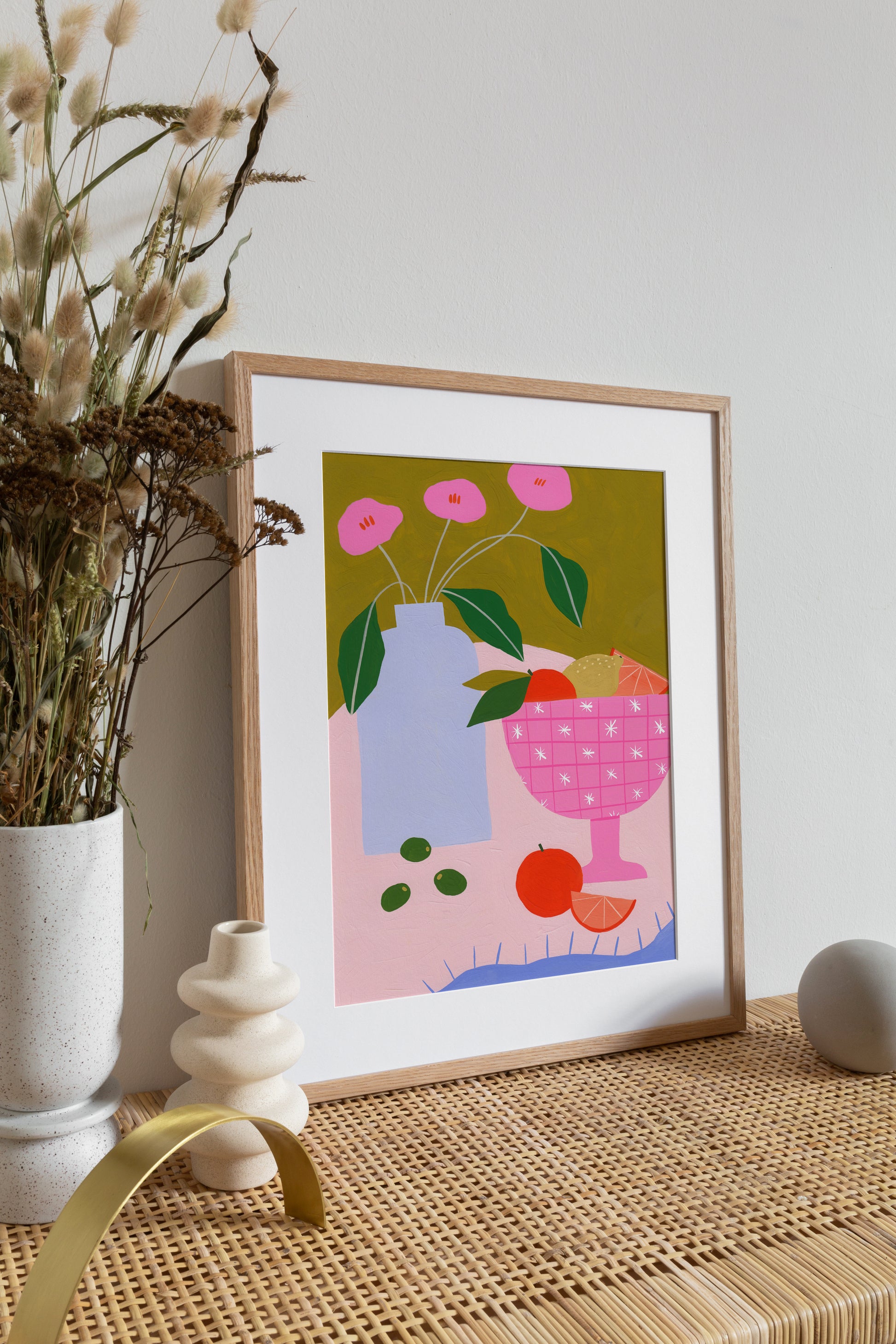 still life print of an original gouache drawing. pink table with a pink bowl of fruit, lemons and oranges. blue vase with pink flowers and an olive green background.